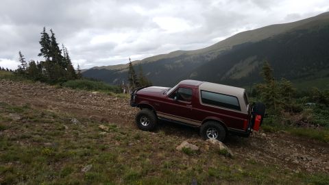 1996 Ford Bronco - A maroon 1996 Ford Bronco is parked on a rocky mountain trail with a scenic view of forested mountains under a cloudy sky. The vehicle has…