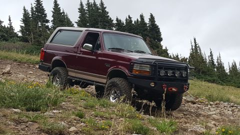 1996 Ford Bronco - A maroon 1996 Ford Bronco is parked on a rocky, grassy hillside with pine trees in the background.