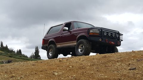 1996 Ford Bronco - A maroon 1996 Ford Bronco with off-road modifications is parked on a dirt hill under a cloudy sky. The vehicle features a custom front bump…