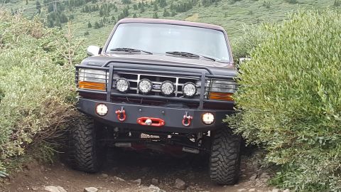 1996 Ford Bronco - The front view of a dark red 1996 Ford Bronco is shown, equipped with a heavy-duty aftermarket bumper featuring multiple KC lights and red…