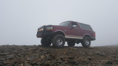 1996 Ford Bronco - A maroon 1996 Ford Bronco with an aftermarket bumper and auxiliary lights is parked on a rocky, uneven terrain with dense fog in the backgr…