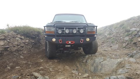 1996 Ford Bronco - A front view of a dark-colored Ford Bronco navigating a rocky, off-road trail with a custom bumper and brush guard. The vehicle is position…
