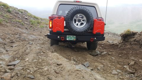 1996 Ford Bronco - The rear of a red and tan Ford Bronco is shown driving on a rocky, off-road trail. It is equipped with a spare tire mounted on the back, tw…