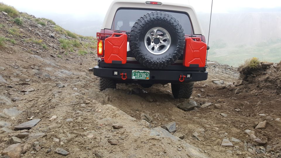 Rear view of a red and tan Ford Bronco on a rocky mountain trail, with a spare tire and jerry cans.
