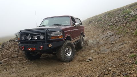 1996 Ford Bronco - A maroon 1996 Ford Bronco is shown from a front 3/4 angle, driving on a rocky, off-road mountain trail with fog in the background. The Bron…