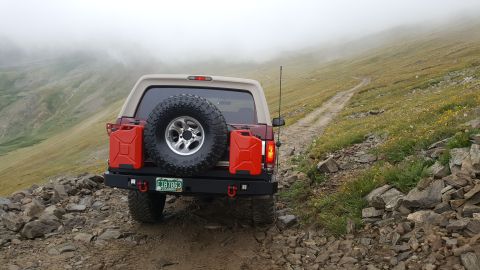 1996 Ford Bronco - The rear of a red 1996 Ford Bronco is shown parked on a rocky mountain trail with two red jerry cans mounted on the back. The vehicle is fa…