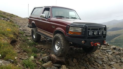 1996 Ford Bronco - A maroon 1996 Ford Bronco is parked on a rocky, mountainous trail with green grass and yellow wildflowers on the left. The vehicle has a bl…