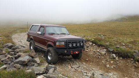 1996 Ford Bronco - A maroon 1990s Ford Bronco with an aftermarket bumper and light bar is parked on a rocky, off-road trail in a foggy, mountainous landscape.