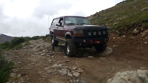 1996 Ford Bronco - A maroon 1990s Ford Bronco is navigating a rocky, uphill trail in a mountainous, cloudy environment. The Bronco features a lifted suspensio…