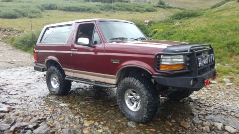 1996 Ford Bronco - A maroon 1996 Ford Bronco with a tan stripe sits partially submerged in a rocky stream bed, with a mountainous, grassy landscape in the bac…