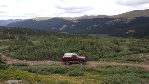 1996 Ford Bronco - A maroon 1996 Ford Bronco is parked on a dirt road in a mountainous, forested landscape.