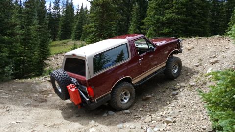 1996 Ford Bronco - A maroon 1996 Ford Bronco with a tan removable top is parked on a rocky, wooded trail. The rear of the vehicle shows a spare tire mounted o…