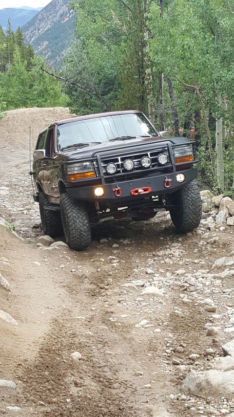 1996 Ford Bronco - A dark red 1996 Ford Bronco is shown from a 3/4 front angle, navigating a rocky, dirt trail in a mountainous, wooded area.