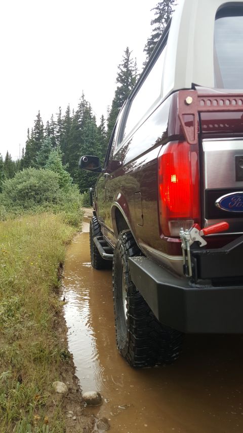 1996 Ford Bronco - The rear passenger side of a maroon Ford Bronco is shown driving through a muddy water puddle on a dirt road, with trees lining the path.