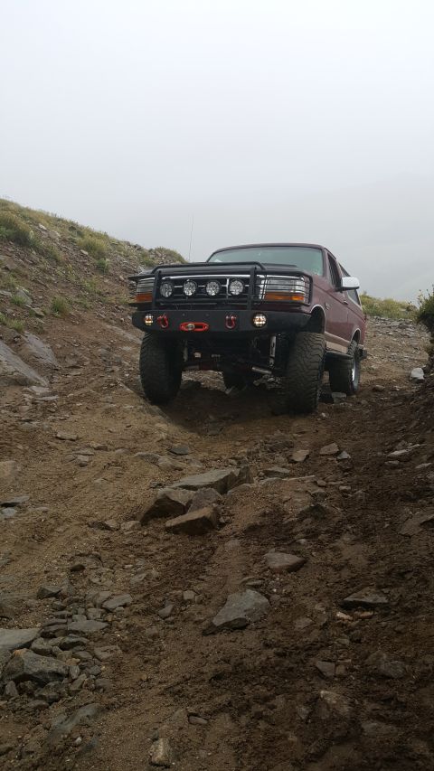 1996 Ford Bronco - A maroon 1996 Ford Bronco is shown from a front 3/4 angle, driving on a rocky, off-road trail with a foggy sky in the background. The Bronc…