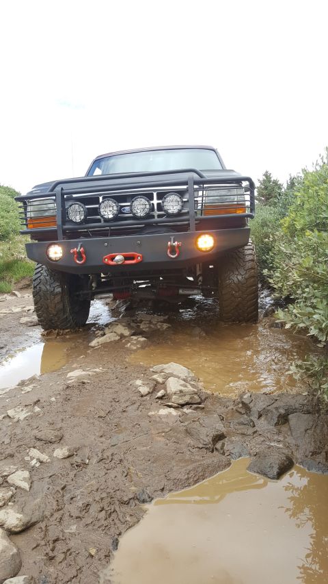 1996 Ford Bronco - A modified black Ford Bronco is driving through a muddy puddle on a rocky trail. The front of the SUV features a heavy-duty bumper with a w…