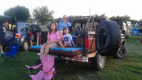 1996 Ford Bronco - A family is gathered around the back of a red Ford Bronco, with two children and a woman sitting in the bed. A blue SUV is parked next to i…
