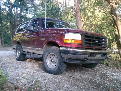 1996 Ford Bronco - A maroon 1996 Ford Bronco is parked on a gravel road surrounded by trees. The front passenger side of the SUV is visible, showing its grill…