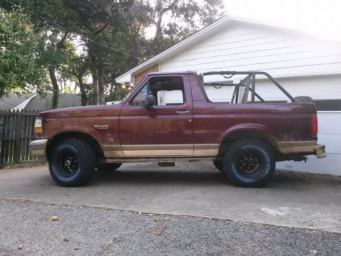 1996 Ford Bronco - A side view of a maroon 1996 Ford Bronco parked on a concrete driveway in front of a white garage. The vehicle has a roll cage installed in…