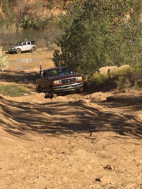 1996 Ford Bronco - A maroon Ford pickup truck with a flag mounted on the back navigates a rough, muddy terrain. A white Jeep SUV is visible in the background…