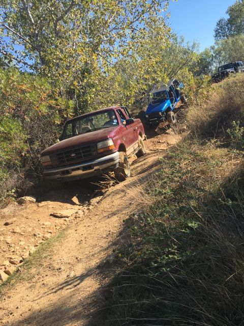 1996 Ford Bronco - A maroon Ford Bronco is driving up a dirt trail with a blue off-road vehicle behind it. The Bronco is angled towards the camera on the left…