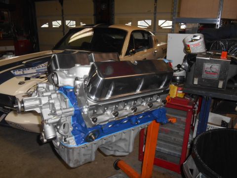 1972 Ford Bronco - A partially assembled V8 engine with chrome valve covers and a blue painted block is shown in the foreground, with the front end of a class…