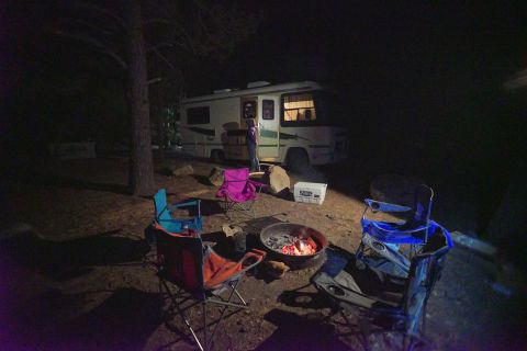 1995 Chevrolet P30 1 Ton - Step Van - A motorhome is parked at a campsite at night, with a campfire burning in the foreground and several camping chairs set up around it.