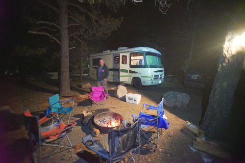 1995 Chevrolet P30 1 Ton - Step Van - A campsite at night features a white and green RV parked next to a dark-colored vehicle. Several camping chairs surround a fire pit in the…