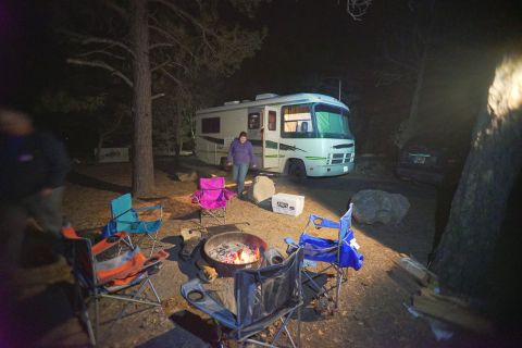 1995 Chevrolet P30 1 Ton - Step Van - green-striped Class A motorhome parked at a forest campsite at night, surrounded by colorful camp chairs and a fire pit.