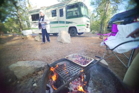 1995 Chevrolet P30 1 Ton - Step Van - A woman stands next to a vintage RV, holding a plate and utensils, while bacon cooks on a grill over a campfire in the foreground.