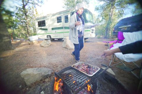 1995 Chevrolet P30 1 Ton - Step Van - A woman in a knitted hat and scarf walks past a motorhome while food is being cooked over a campfire grill. The motorhome is white with gre…