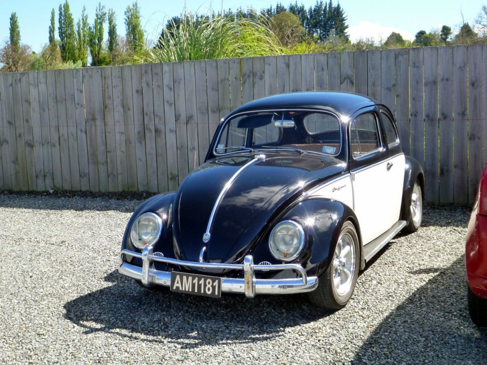 Black and white two-door Volkswagen Beetle with chrome trim and license plate AM1181 parked on gravel.