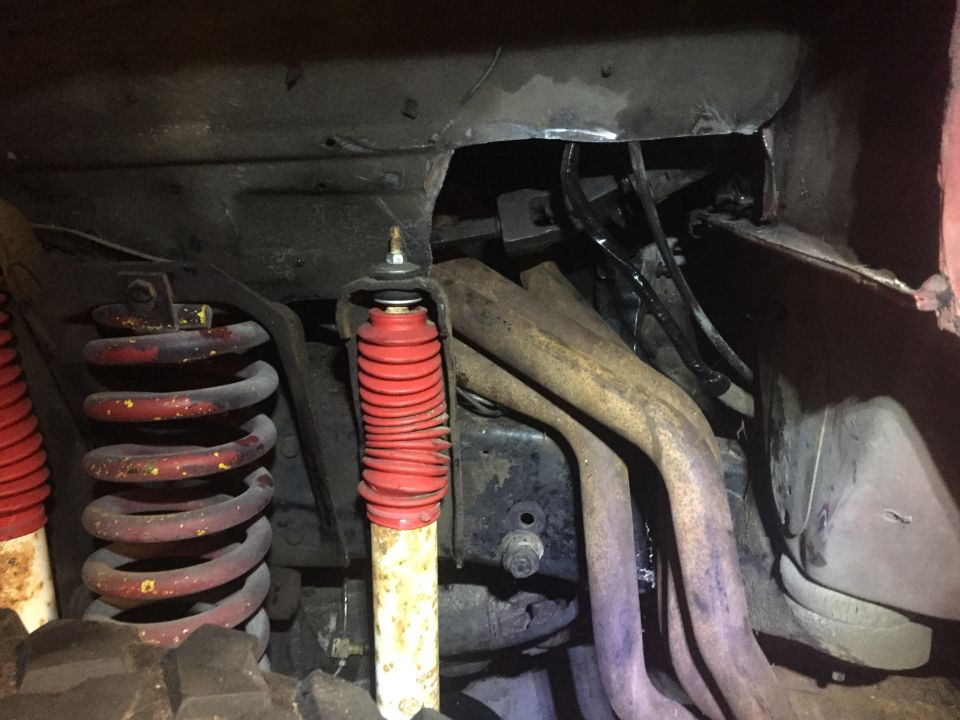 Underside of a vehicle showing red coil springs, shock absorber with red boot, and exhaust pipes.