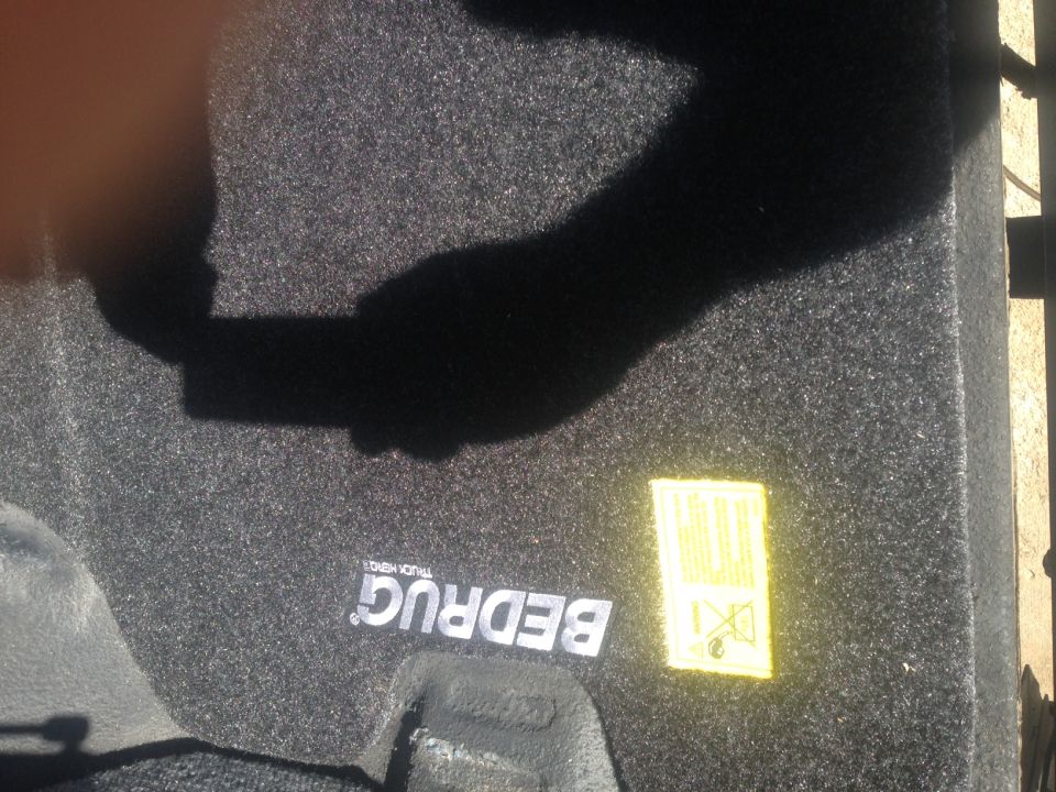 Black Bedrug truck bed liner with yellow warning sticker and shadow of hand holding phone.