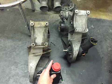 1988 Ford Bronco - Two power steering pump brackets are displayed on a dark gray surface. The bracket on the right includes the power steering pump and reserv…