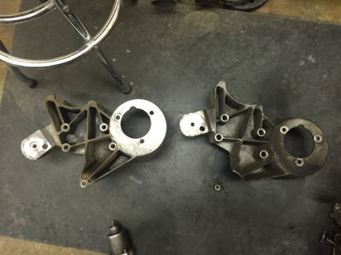 1988 Ford Bronco - Two aluminum engine brackets are laid out on a dark, textured surface. The bracket on the left has a circular flange with three bolt holes,…