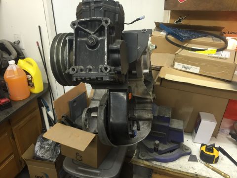 1988 Ford Bronco - A black engine component with a large pulley is mounted in a vise on a workbench. Various boxes, bottles of liquid, and tools are scattered…