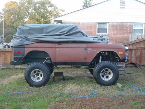 1972 Ford Bronco - A modified 1972 Ford Bronco is covered with a tarp and parked on a grassy lawn in front of a brick house. The vehicle features large off-ro…