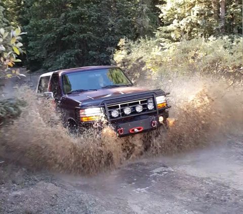 1996 Ford Bronco - A dark colored Ford Bronco is driving through a muddy puddle on a dirt road surrounded by trees. The vehicle has a black grille guard with…