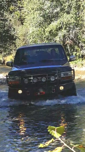 1996 Ford Bronco - A dark colored Ford Bronco is driving through shallow water with trees in the background. The vehicle has a brush guard with multiple auxil…