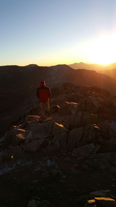 1996 Ford Bronco - A person stands on a rocky mountain summit, looking out at a sunset over a range of mountains. The sky is clear and blue, transitioning to…