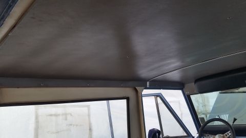 1973 Ford Bronco - The interior ceiling of a vehicle is shown, with a dark gray headliner attached to metal framing. Visible through the windows are parts of…