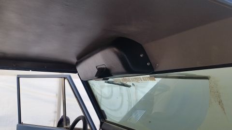 1973 Ford Bronco - A black overhead storage console is mounted to the headliner of the vehicle, above the windshield. The interior of the vehicle is visible,…