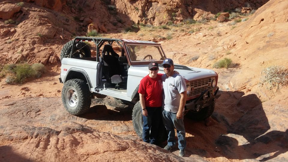Couple posing in front of a light gray Ford Bronco with off-road tires and a roll cage in a desert.