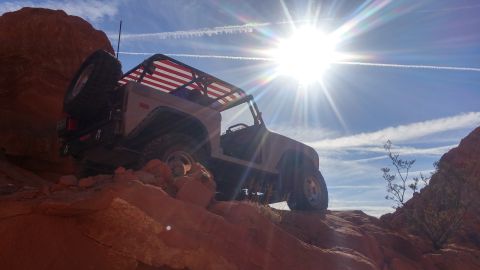 1979 Ford Bronco - A tan 1979 Ford Bronco is parked on a rocky, desert terrain under a bright sun and blue sky with contrails. The vehicle has an American fla…