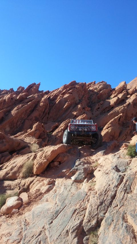 1979 Ford Bronco - A red 1979 Ford Bronco is navigating a steep, rocky off-road trail under a clear blue sky. The vehicle is equipped with large tires and off…