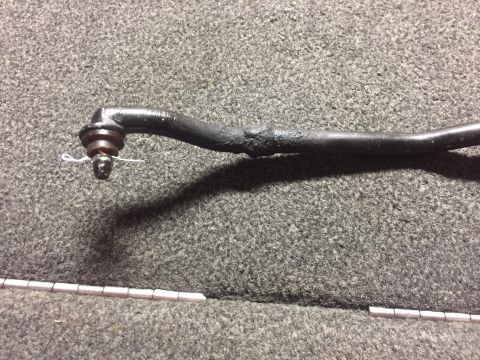 1982 Ford Bronco - A close-up view of a tie rod end with a cotter pin. The part is dark colored and appears to be made of metal, resting on a textured gray su…