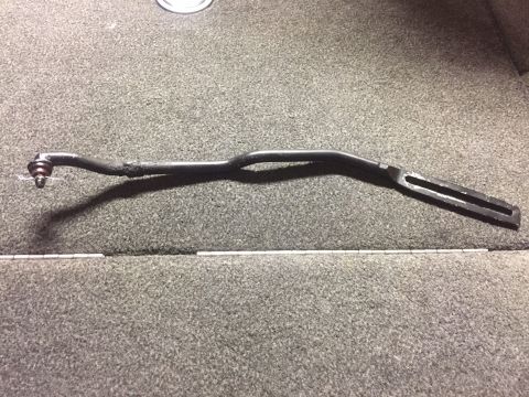 1982 Ford Bronco - A black tie rod end with a ball joint is shown attached to a bent metal rod. The rod has a slotted end and is lying on a grey carpeted surf…