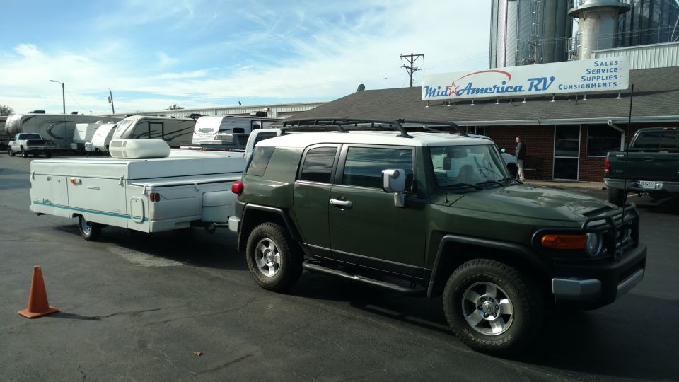 Dark green Toyota FJ Cruiser towing a white pop-up camper in front of an RV dealership.