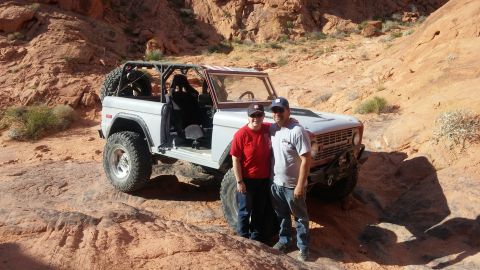 1979 Ford Bronco - A man and a woman pose in front of a light gray Ford Bronco with a roll cage in a rocky, desert landscape.